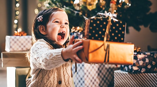 Toddler girl smiles and shows a missing tooth as she holds a stack of gift-wrapped packages.